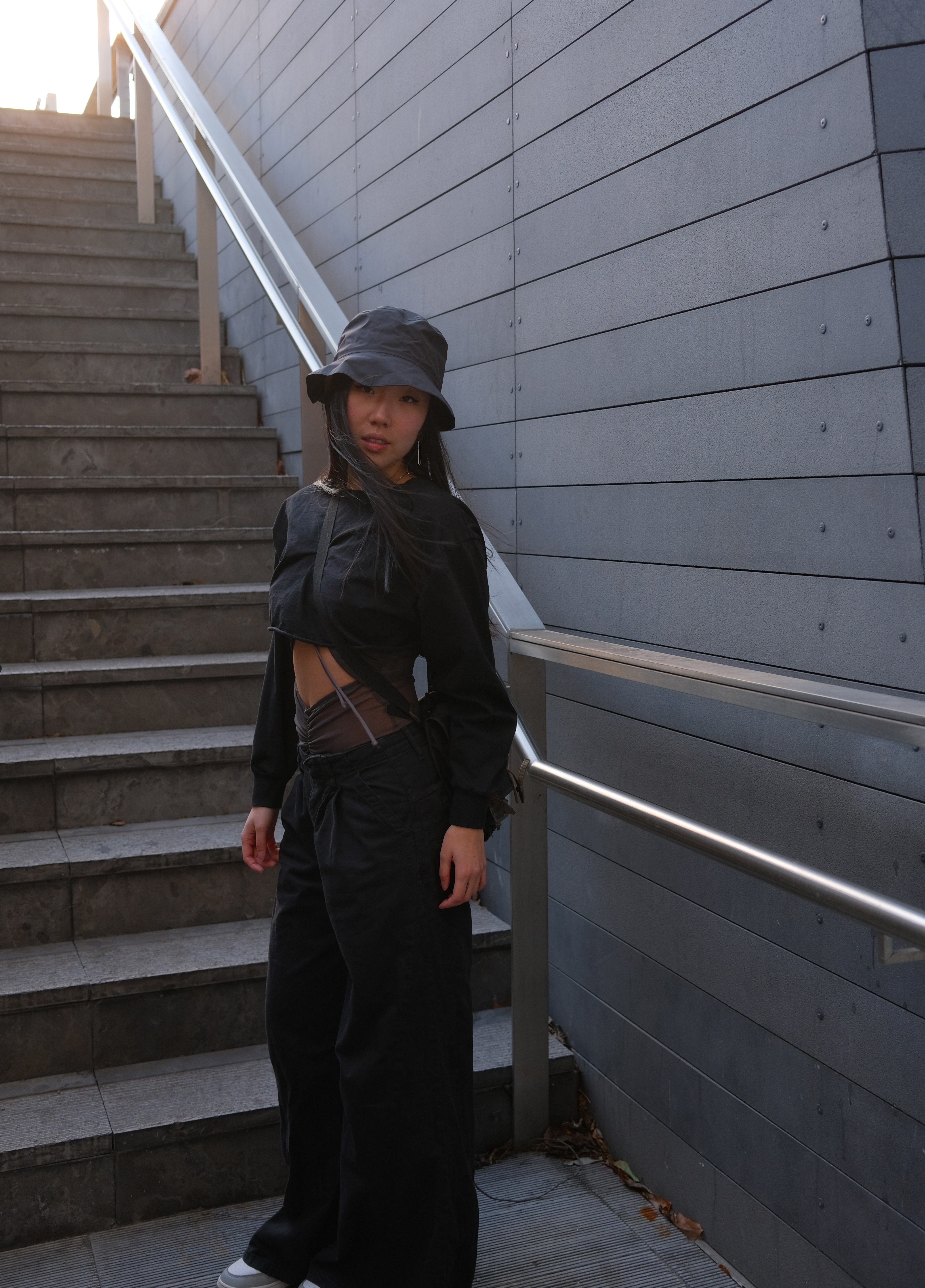 Girl standing at the bottom of the starcaise wearing a bucket hat, longsleeve black crop top, and baggy pants. The girl is asian, has long brown hair, and is wearing smoke grey Jordans